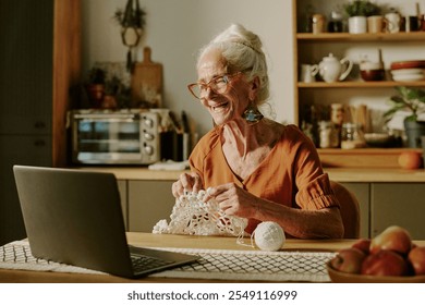 Elderly woman with glasses seated at wooden table working on crochet project while smiling and using laptop in inviting home kitchen with warm lighting - Powered by Shutterstock