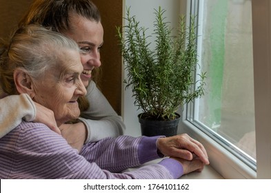 An elderly woman, a grandmother looks out the window, a granddaughter approaches her and hugs, a happy family - Powered by Shutterstock