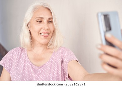 Elderly woman with gray hair taking a selfie with smartphone while smiling in relaxed casual outfit indoors. - Powered by Shutterstock