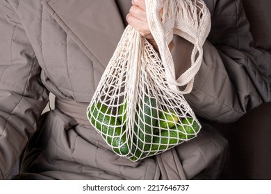 An Elderly Woman In A Green Quilted Coat Holds A String Bag With Cucumbers In Her Hand