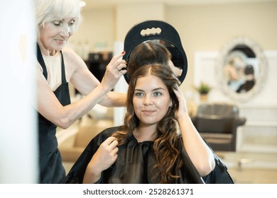 Elderly woman hairdresser holds mirror behind girl client and shows result of work. Surprised and delighted, client rejoices with new haircut - Powered by Shutterstock