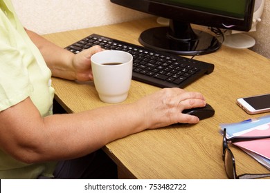 An Elderly Woman Hand On A Computer Mouse. The Old Grandmother Works Behind The Computer Keyboard And Drinks Coffee.