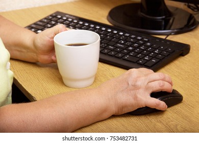 An Elderly Woman Hand On A Computer Mouse. The Old Grandmother Works Behind The Computer Keyboard And Drinks Coffee.