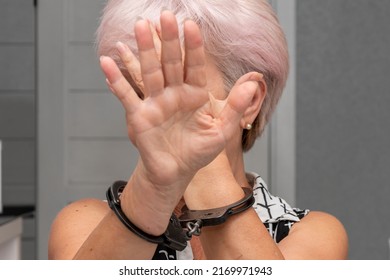 An Elderly Woman In Handcuffs Hides Her Face On A Blurry Background. Elderly Criminal, Criminal Liability Of Pensioners, Domestic Violence Against Women.