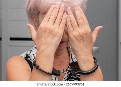 An Elderly Woman In Handcuffs Hides Her Face On A Blurry Background. Elderly Criminal, Criminal Liability Of Pensioners, Domestic Violence Against Women.