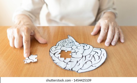 Elderly woman hands putting missing white jigsaw puzzle piece down into the place as a human brain shape. Creative idea for memory loss, dementia, Alzheimer's disease and mental health concept.