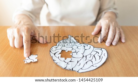 Elderly woman hands putting missing white jigsaw puzzle piece down into the place as a human brain shape. Creative idea for memory loss, dementia, Alzheimer's disease and mental health concept.