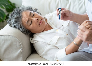 Elderly woman having cold during lie down on couch while Senior man checking temperature of his wife with digital thermometer in house. Old female having fever. Illness, Disease, virus. BeH3althy - Powered by Shutterstock