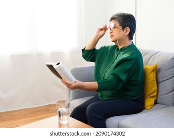Elderly woman having troubles with long eye-sight trying to read a book, sitting on sofa at home.