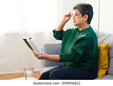 Elderly woman having troubles with long eye-sight trying to read a book, sitting on sofa at home.