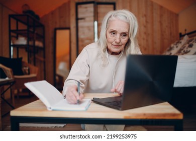 Elderly Woman With Headphones Talking Video Call On Laptop In Kitchen, Waving At Screen, Chatting And Speaking Camera Device View.