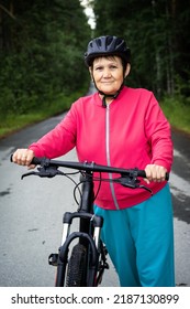 Elderly woman in helmet stands next to bicycle on forest road. Senior lady doing cycling in wooded place. Healthy, sportive, active lifestyle in old age