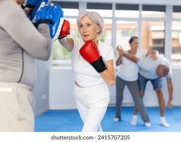 Elderly woman with her trainer at gym. Woman wearing boxing gloves exercise and punch to pads for boxing - Powered by Shutterstock