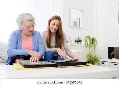 Elderly Woman With Her Young Granddaughter At Home Looking At Memory In Family Photo Album