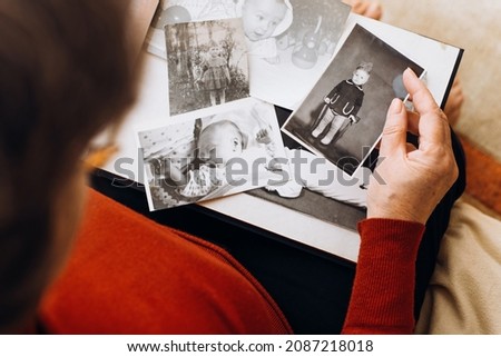 An elderly woman holding album with black and white retro photographs of childhood, remembering herself as little girl, child. Happy memories, nostalgia concept. Selective focus on vintage, old images