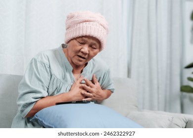 Elderly woman holding chest in pain, sitting on couch. Wearing pink beanie, indicating cancer treatment. Expression of discomfort, vulnerability. conveys health struggles, need for care and empathy. - Powered by Shutterstock