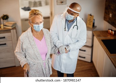 Elderly Woman Holding Hands With African American Doctor While Using Walking Cane At Nursing Home. They Are Wearing Face Masks Due To Coronavirus Pandemic. 