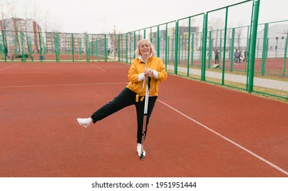 An Elderly Woman Holding A Nordic Walking Stick Stands At The Stadium On A Red Treadmill. Healthy Lifestyle Of Old People