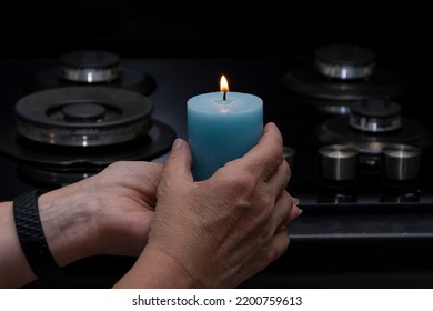 An Elderly Woman Holds A Burning Candle In Her Hands, Against The Background Of A Non-working Gas Stove, Selective Focus. Concept: Turning Off Heating And Gas, Reducing Room Temperature.