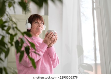 elderly woman listening to music or a podcast by a window, reflecting on the complexities of aging and the impact it has on our emotional wellbeing. - Shutterstock ID 2290306579