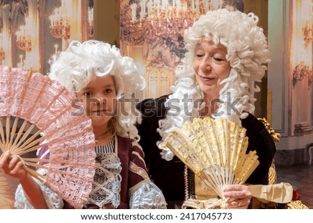 An elderly woman and a little girl dressed in elegant 18th century style clothing pose with hand fans at a costumed historical event, 