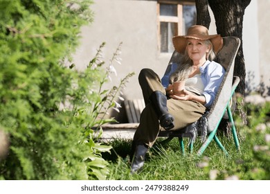  elderly woman  with long grey hair wearing a straw hat sits in a garden wheelbarrow and drinks tea in the summer garden near the house. - Powered by Shutterstock