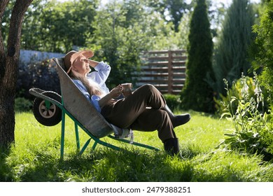  elderly woman  with long grey hair wearing a straw hat sits in a garden wheelbarrow and drinks tea in the summer garden near the house. - Powered by Shutterstock