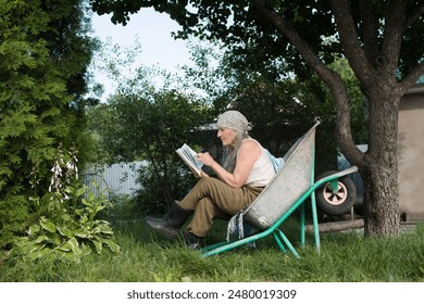  elderly woman  with long grey hair  sits in a garden wheelbarrow and and reads a book in the summer garden near the house.  - Powered by Shutterstock