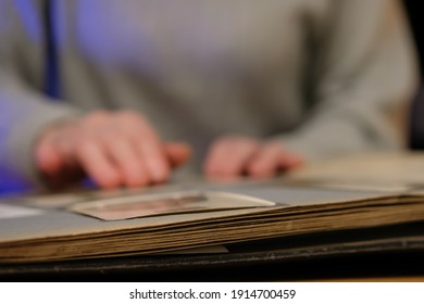 Elderly Woman Looks Through An Family Album With Old Photos At Table At Home. Old Granny Memories Past Times And Remembering His Life.