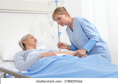 Elderly Woman Lying In The Hospital Room Bed, Nurse Holding The Needle To Connect The IV To The Patient's Arm
