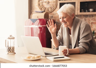 Elderly woman making video call on laptop in kitchen, waving at screen, chatting with children, free space - Powered by Shutterstock