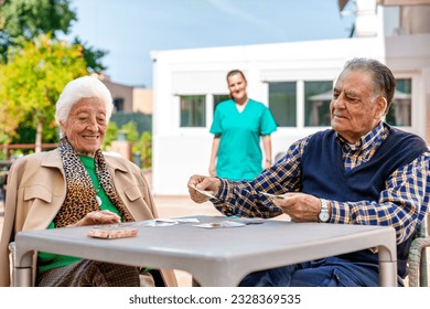 An elderly woman and man sit at a patio table in a nursing home, engrossed in a card game. Bathed in sunlight, a beautiful garden surrounds them as a nurse approaches, adding to their joy. - Powered by Shutterstock