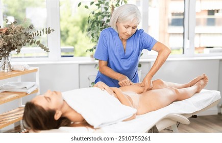 Elderly woman masseuse doing thigh massage to young woman client in massage room - Powered by Shutterstock