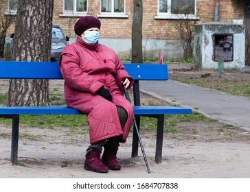 An Elderly Woman In A Medical Mask Sits On A Bench Alone.  Coronavirus Epidemic, Self-isolation . Kiev Ukraine March 27, 2020