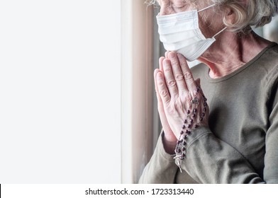 Elderly Woman In A Medical Respiratory Mask Near The Window Prays With A Rosary And A Cross. Religion. Prayer For Human Health. The Epidemic Of Coronavirus Covid-19. Quarantine And Isolation.Copyspace