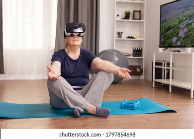 Elderly Woman Meditating With VR Headset In The Middle Of The Room. Old Person Pensioner Online Internet Exercise Training At Home Sport Activity With Dumbbell, Resistance Band, Swiss Ball And VR