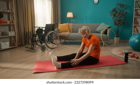 An elderly woman with mobility impairment sits on the floor in the living room next to a wheelchair. A grayhaired woman gives herself a foot massage. The concept of physical rehabilitation. - Powered by Shutterstock