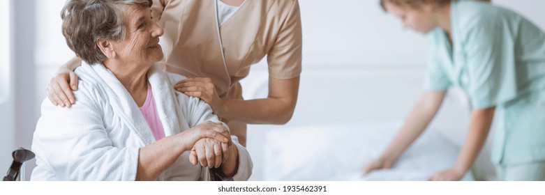 Elderly woman on wheelchair in nursing home with helpful doctor at her side and young nurse making the bed