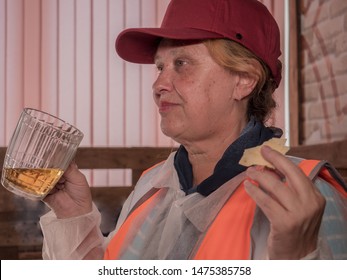 
Elderly Woman In Orange T-shirt And Helmet Tastes Beer At Tasting In Brewery. Photography On Background Of Filling Cranes, Equipment For The Production Of Beer