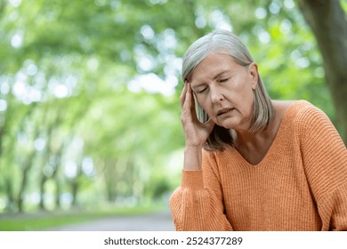 Elderly woman outdoors with closed eyes, holding head, showing stress and headache. Wearing orange sweater, standing in green park. Captures concept of discomfort, pain, and stress among seniors. - Powered by Shutterstock