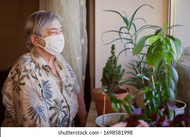 An Elderly Woman A Pensioner Sits At Home Alone In Quarantine In A Protective Mask. Coronavirus Pandemic Covid 19. Social Isolation Distance Looks Out The Window. Upset Grandmother Age. Depression