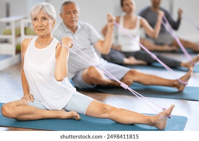 Elderly woman performs weighted exercises during group Pilates workout, stretches elastic band by hand foot while sitting on mat. Concept of active lifestyle, maintaining physical fitness slimness - Powered by Shutterstock