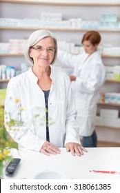 Elderly Woman Pharmacist In Her Pharmacy Standing At The Counter In Her White Lab Coat As A Female Assistant Checks Stock On The Shelves Behind Her