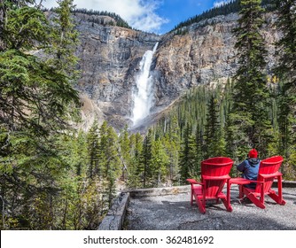 Elderly Woman Photographs The Famous Waterfall. Two Red Deck Chairs To Relax In Front Of The Waterfall Takakkaw. Autumn Day In Yoho National Park In The Rocky Mountains 