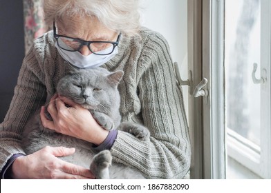 Elderly woman in protective mask holding a cat looks out the window wearing. Christmas quarantine covid19. Coronavirus epidemic. Waiting. Depression. Insulation at home. Pets save you from loneliness