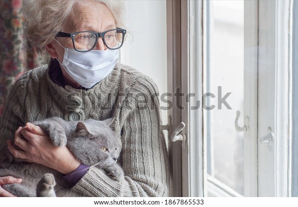 Elderly woman in protective mask holding a cat\
looks out the window wearing. Christmas quarantine covid19.\
Coronavirus epidemic. Waiting. Depression. Insulation at home. Pets\
save you from\
loneliness