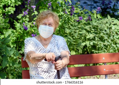 An Elderly Woman In A Protective Medical Mask Is Sitting On A Park Bench.