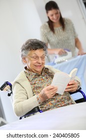 Elderly Woman Reading Book While Home Helper Irons Laundry