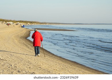 An elderly woman in a red jacket with walking stick walks along the sandy Baltic seashore - Powered by Shutterstock