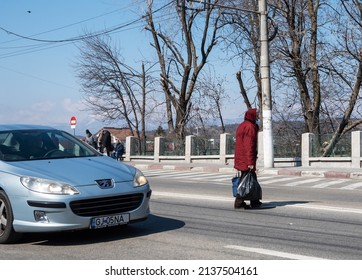 An Elderly Woman Returns From The Market And Crosses The Street.The Old Woman Crosses The Street Among The Cars - She Does Not Cross The Pedestrian Crossing.Romania, Gorj.March,18,2022